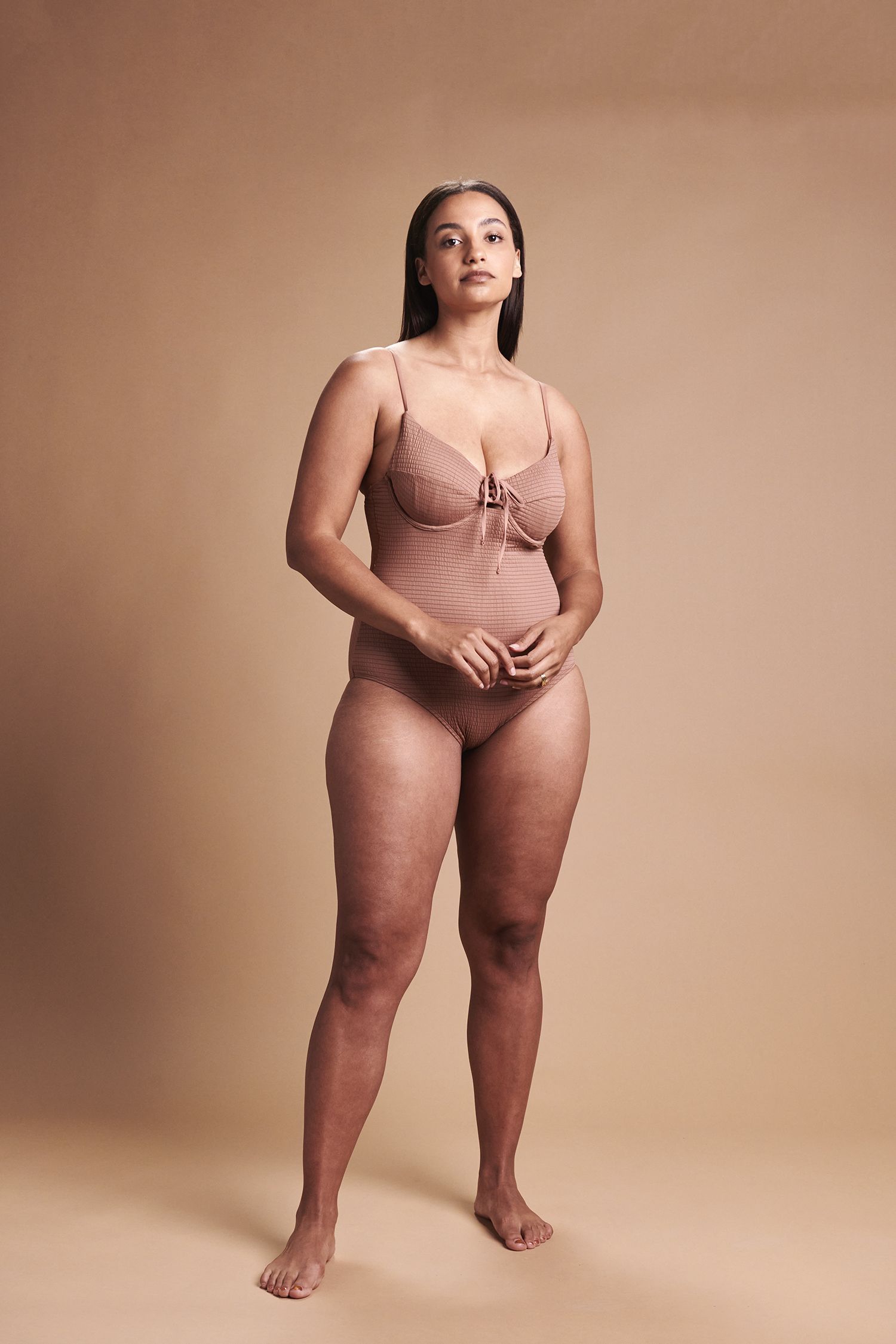 A woman in lingerie is standing in front of a brown background.