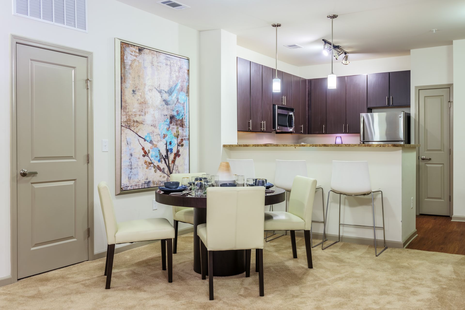 Dining area with round table, four chairs, and kitchen in the background. Beige carpet and a large abstract painting.