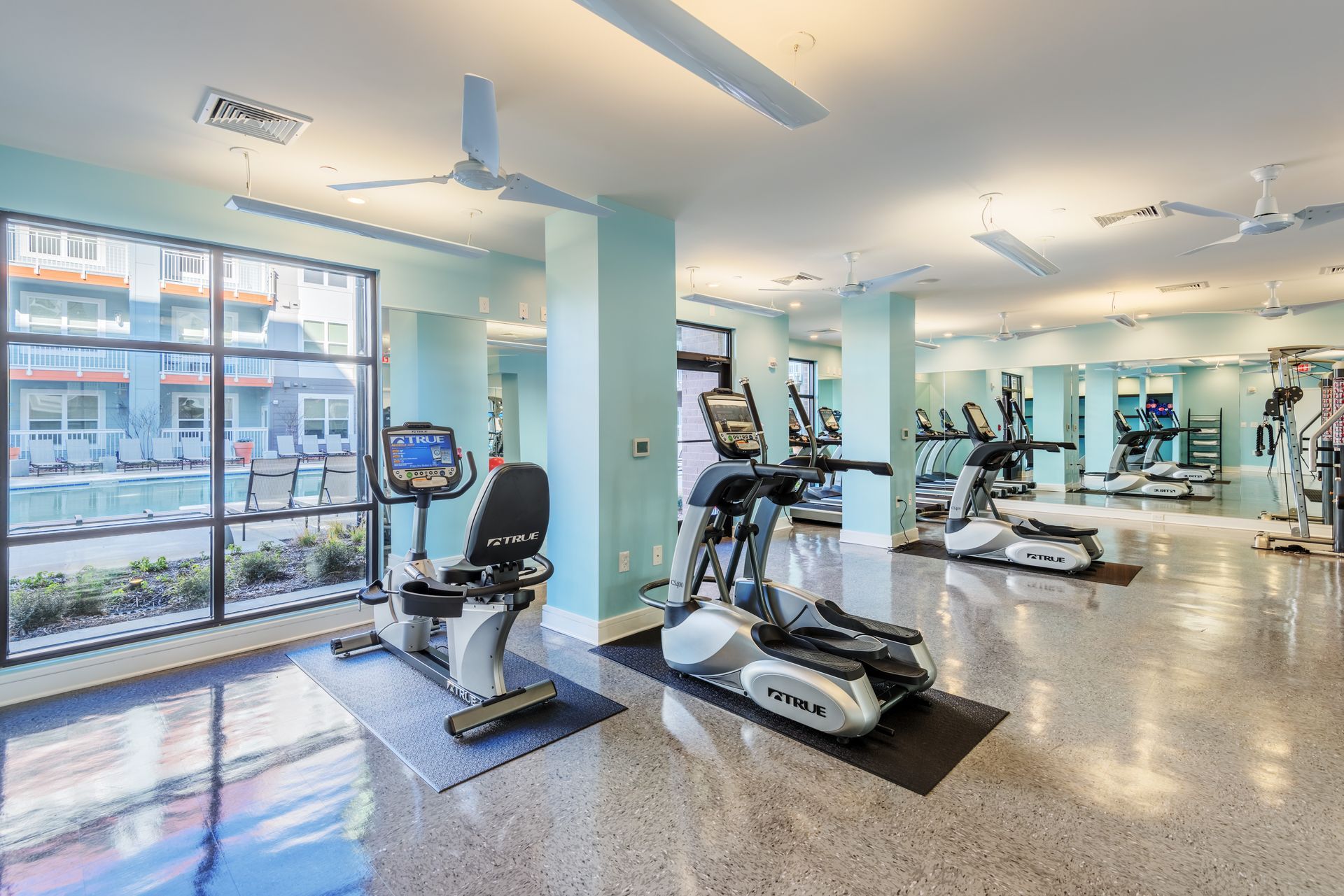 Gym with exercise machines, large windows, and reflective floor.