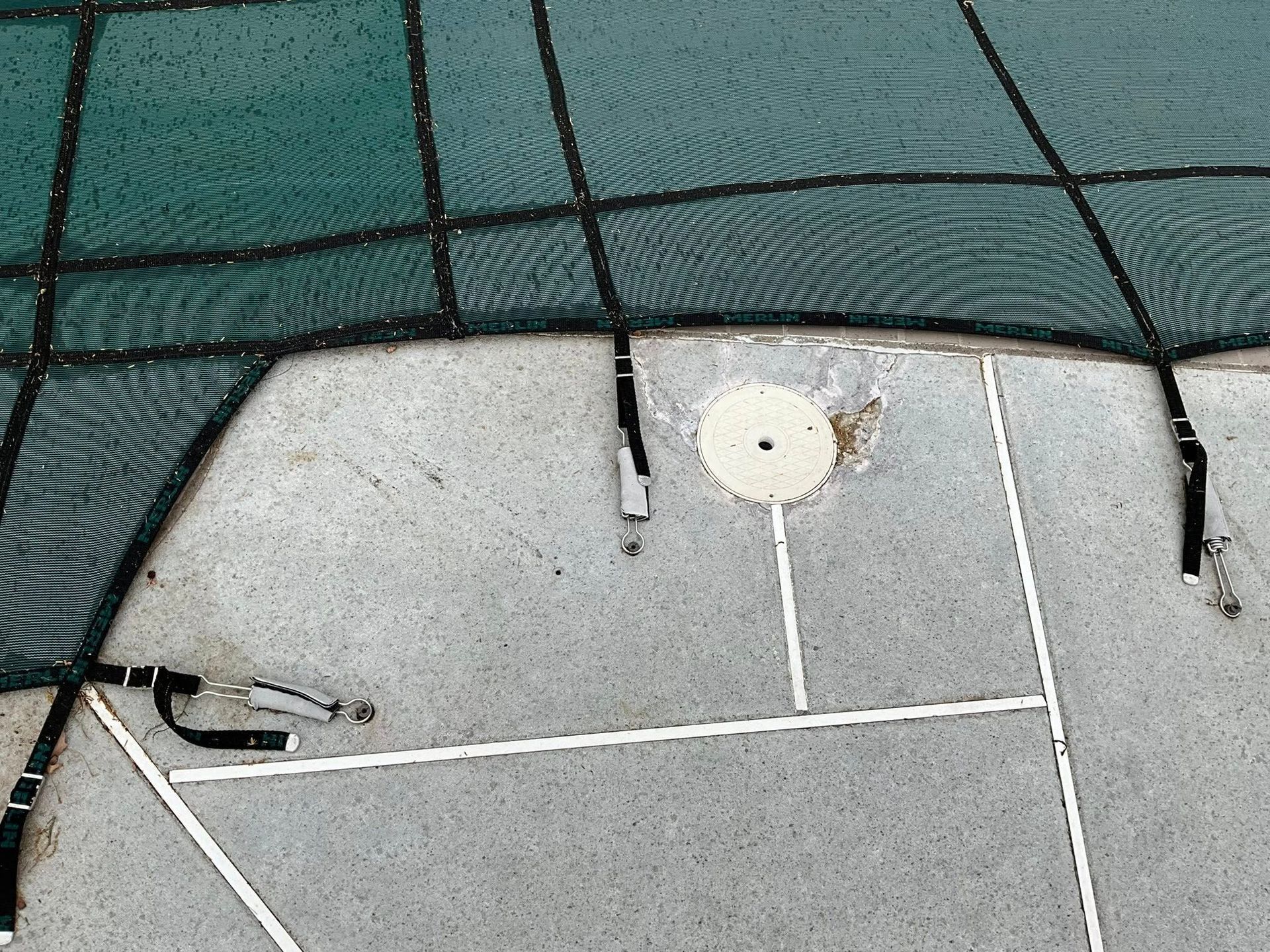 Partial view of a turquoise net over a concrete court with painted white lines and a circular drain