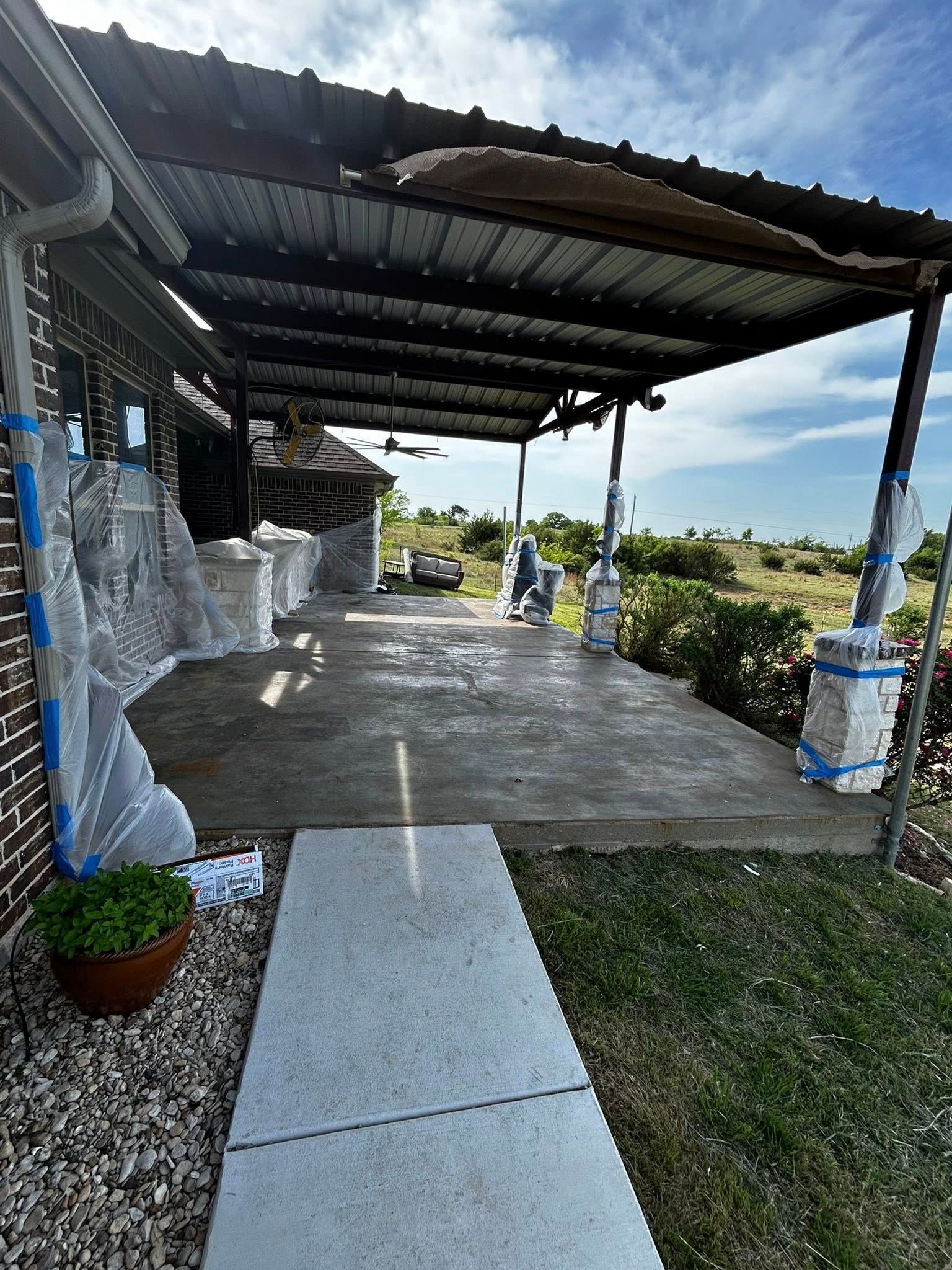 Concrete patio with metal awning, covered in plastic, with a pathway leading towards a garden.