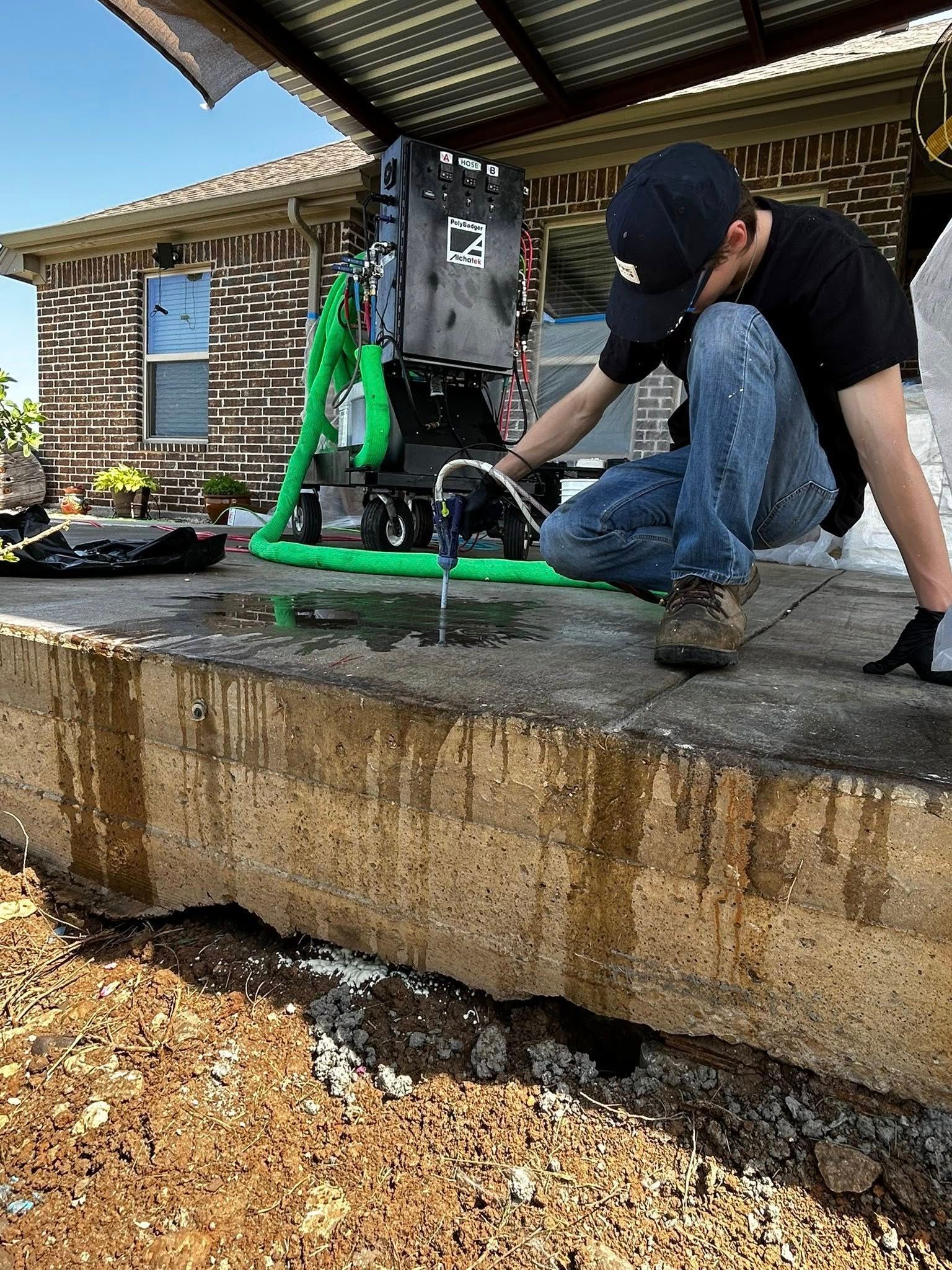 Man drilling concrete with a machine, near a house. Ground is visible, with green tubing.