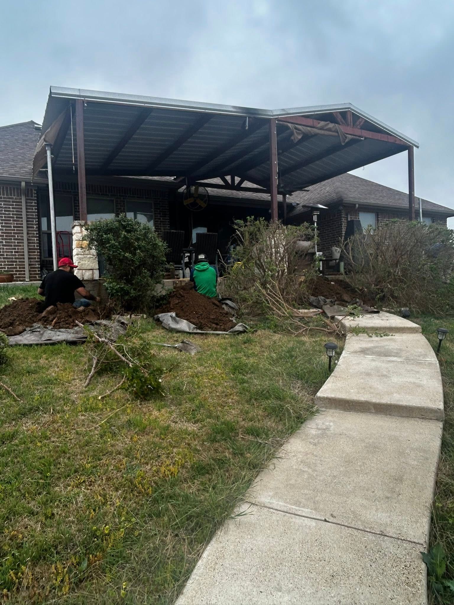 Workers landscaping near a home with a covered patio. Concrete walkway leads to patio. Overcast day.
