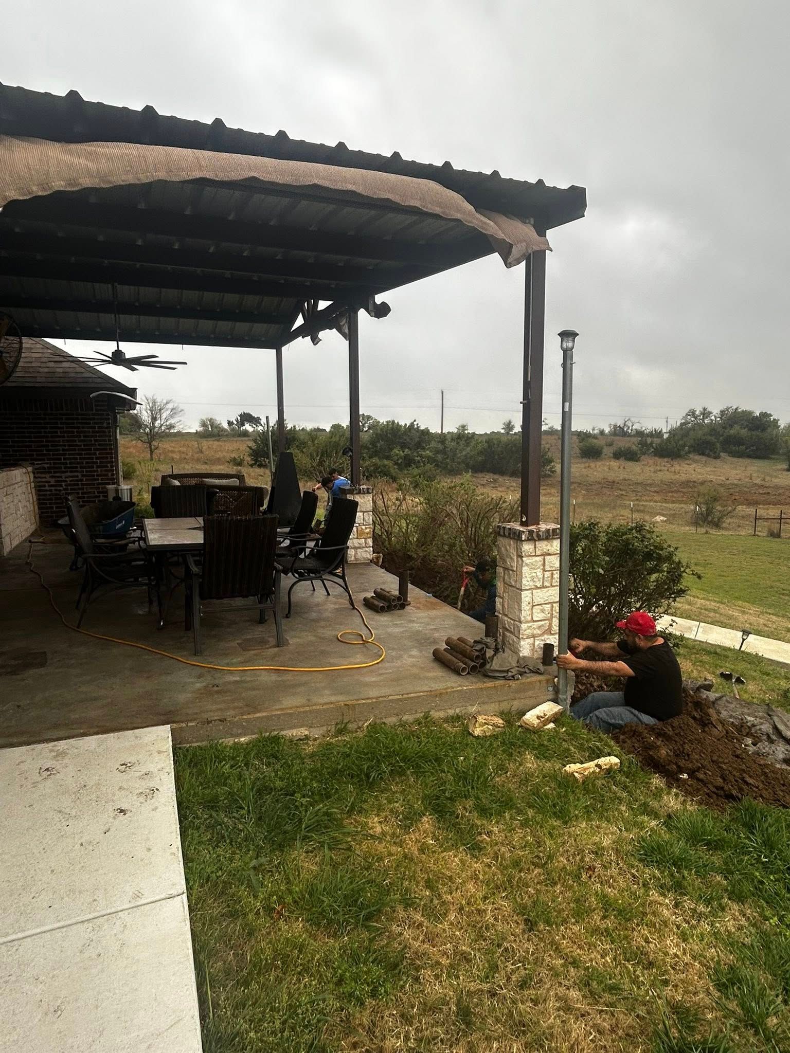 Man planting a post near a patio with a table and chairs under a covered area on a cloudy day.