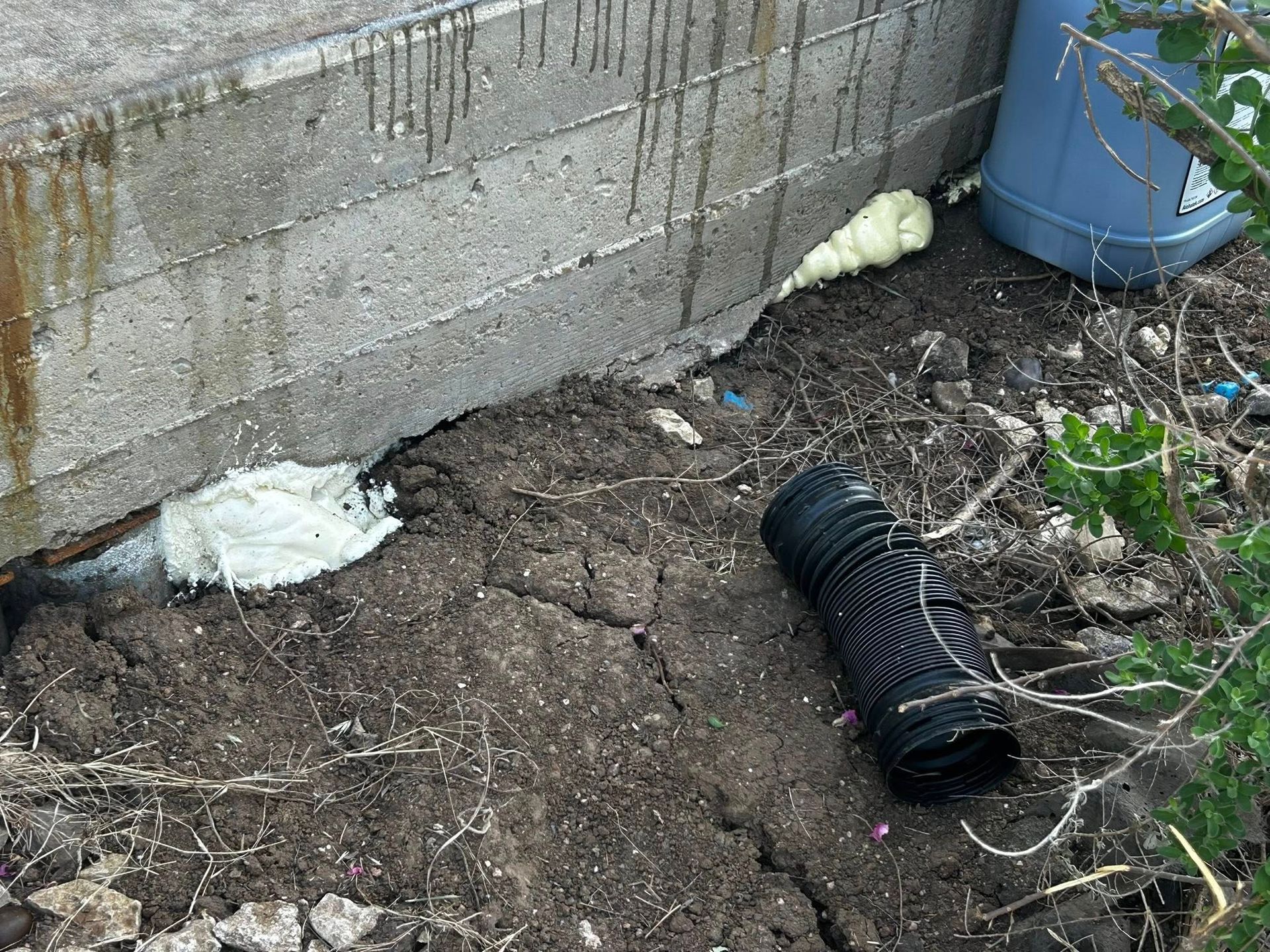 A concrete wall with spray foam sealant, cracked soil, and a black drainage pipe.