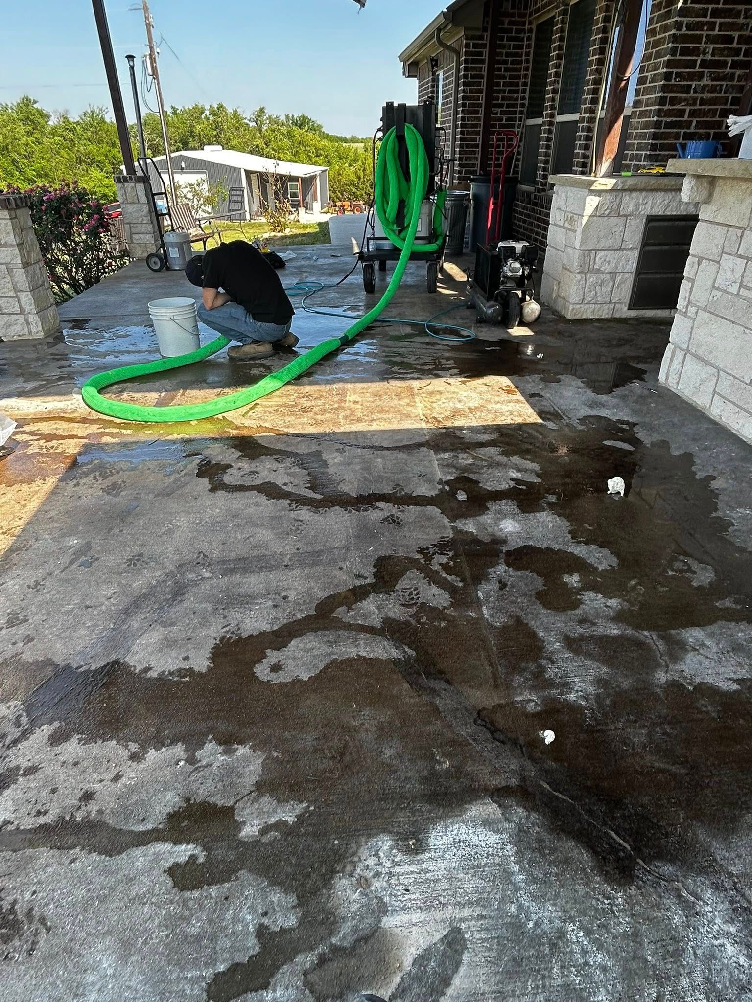 Person kneeling, washing concrete patio with a green hose; sunny outdoor setting.