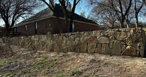 Stone retaining wall in front of a brick house, with bare trees and a blue sky.
