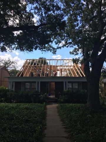 House undergoing roof construction; workers on the wooden frame. Green lawn and trees in front. Sunny, blue sky.