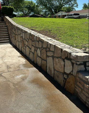 Stone retaining wall next to a concrete driveway and stairs leading up to a grassy lawn.
