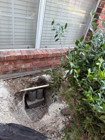 Excavated area near a brick home's window. Exposed concrete piers and a small bush.