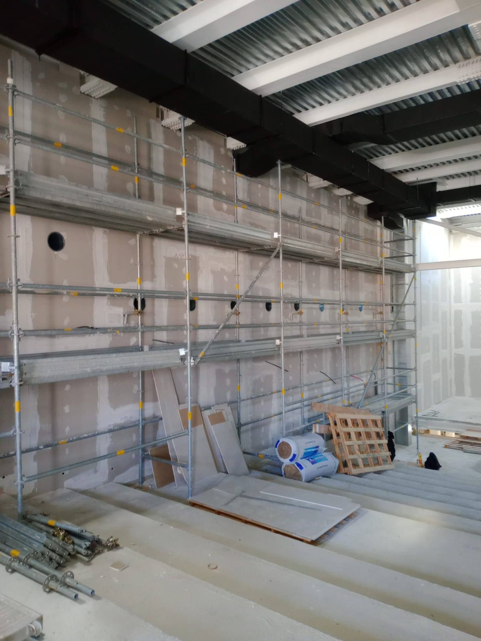 Cantiere: impalcature contro un muro grezzo, gradini in cemento, condotti a soffitto a vista, materiali da costruzione.