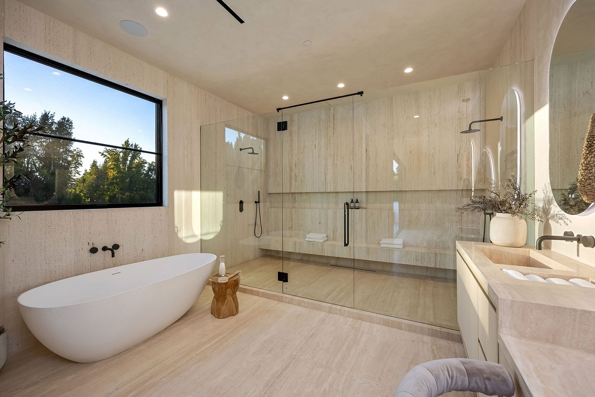 Spacious modern beige bathroom with a freestanding tub, walk-in shower, and large window overlooking trees