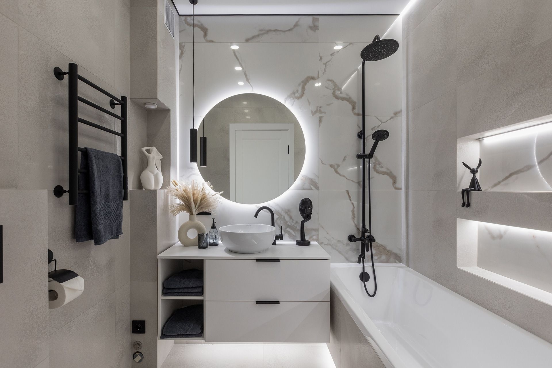 Modern bathroom with a marble wall, round backlit mirror, white vanity, and black hardware fixtures over a white bathtub.