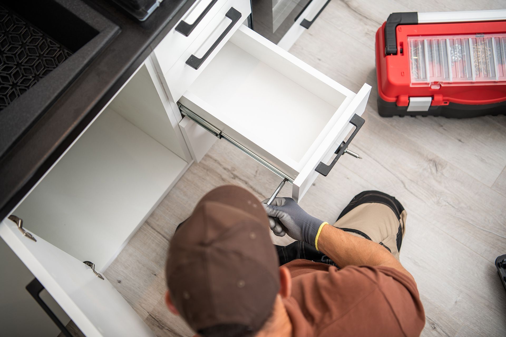 A worker wearing a brown cap and gloves installs a drawer in a kitchen cabinet, with a toolbox nearby on the floor.