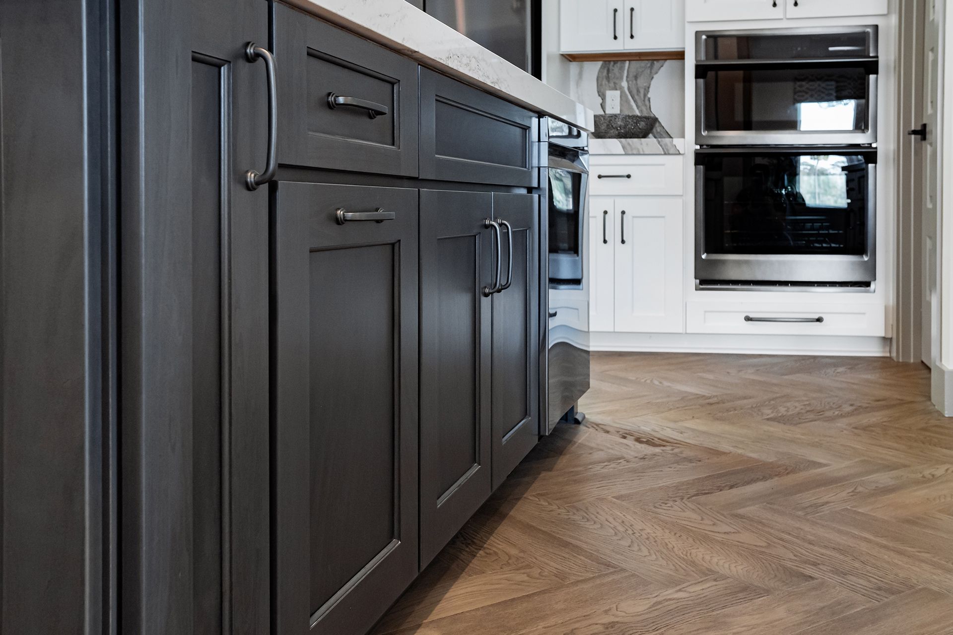 A kitchen interior with dark gray cabinets, white upper cabinets, a stainless steel wall oven, and herringbone wood floors.