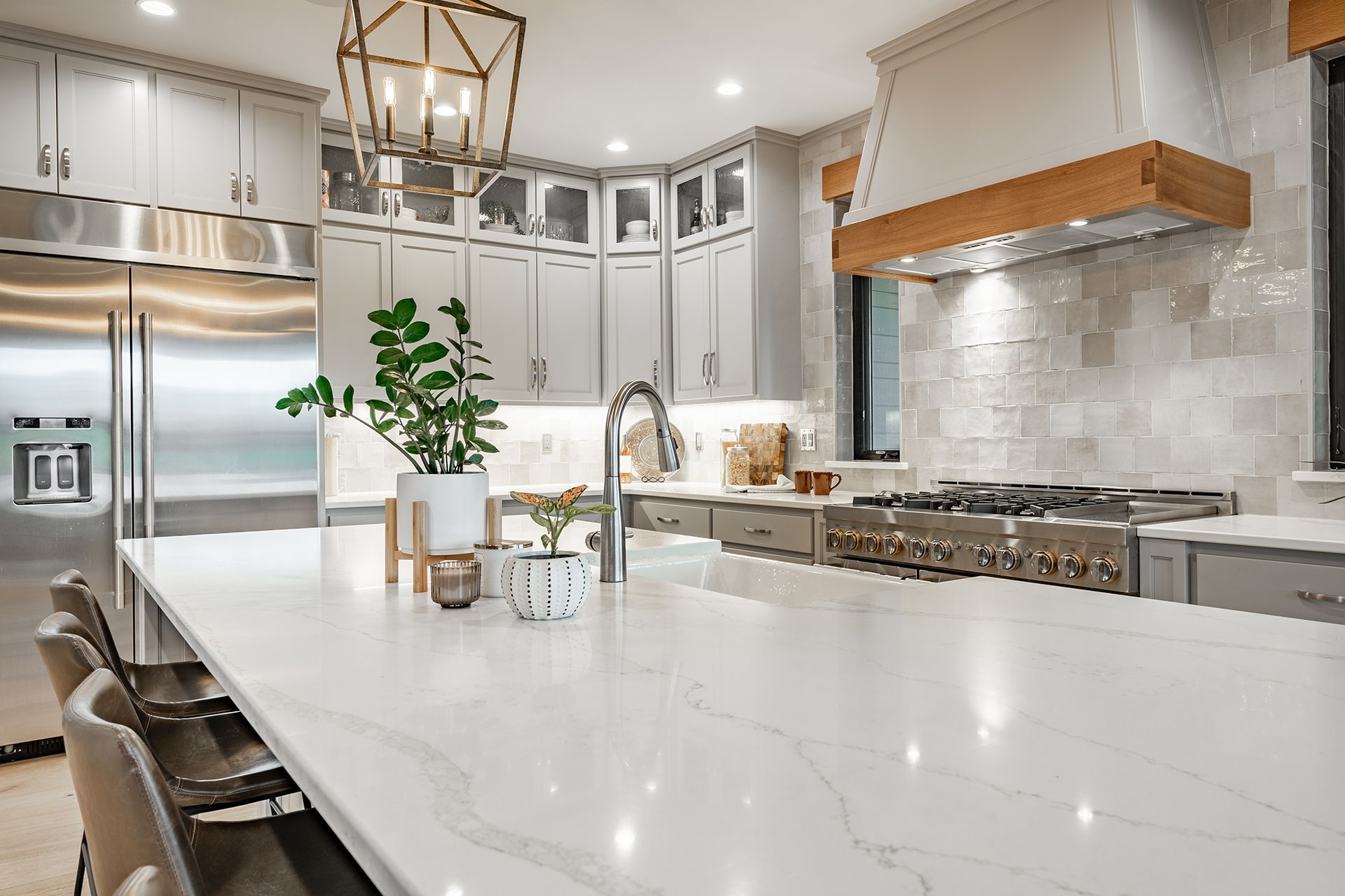 A modern kitchen with a large white quartz island, gray cabinets, stainless steel appliances, and a wood-accented hood.
