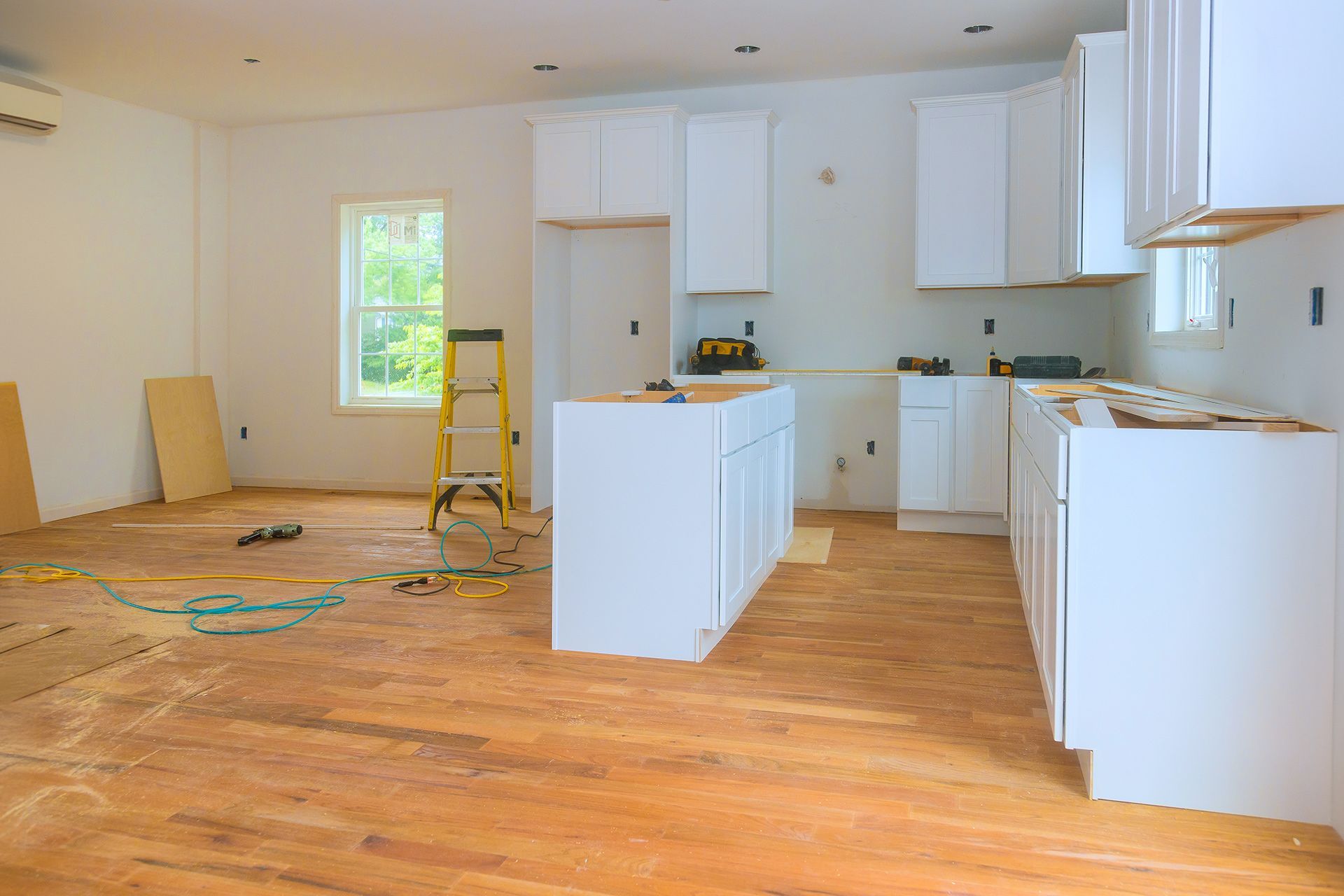 A kitchen under construction featuring white cabinets, a central island, and a yellow stepladder on a wooden floor.