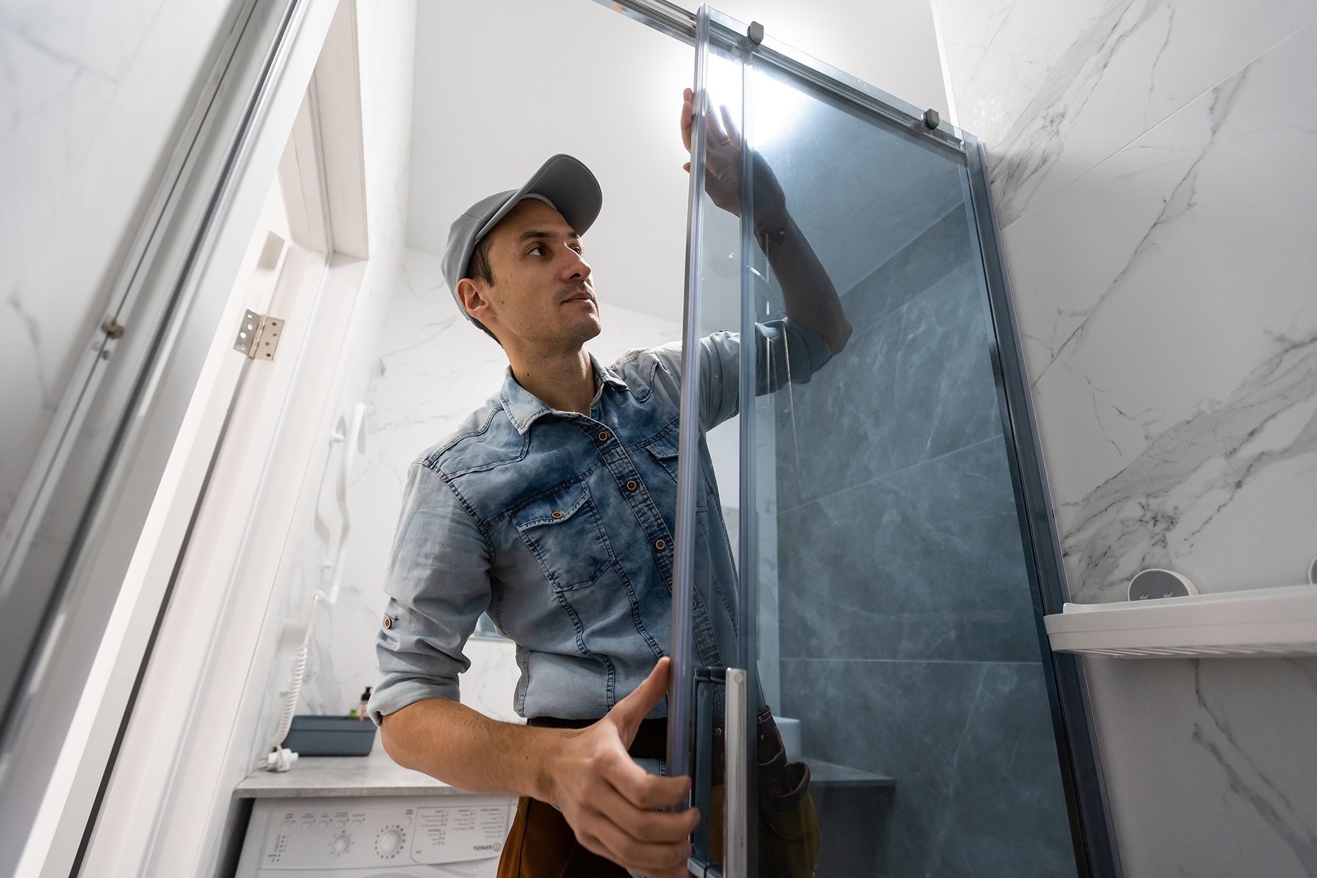 A worker in a cap and denim shirt installs a glass shower door in a modern bathroom.
