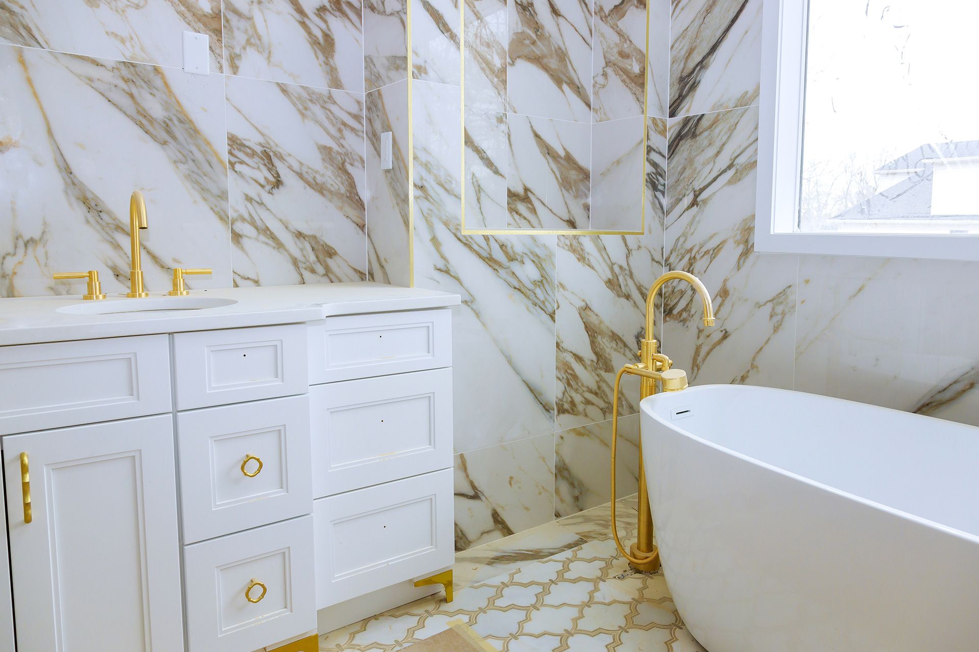 Luxury bathroom with white marble walls, gold fixtures, a freestanding soaking tub, and a white vanity with gold hardware.