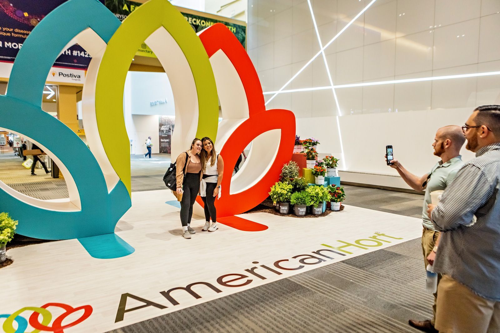 a man taking a picture of two women in front of the AmericanHort logo