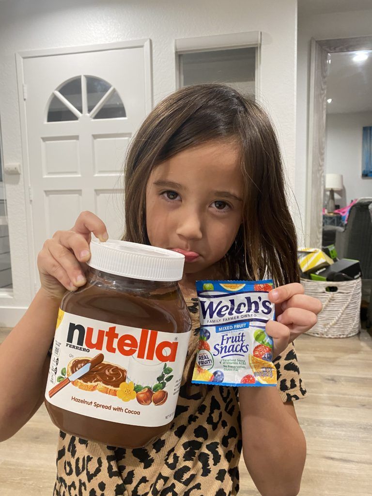 A little girl is holding a jar of nutella and a bag of welch 's fruit snacks.