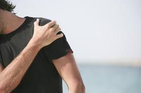 A man is holding his shoulder in pain while standing on the beach.