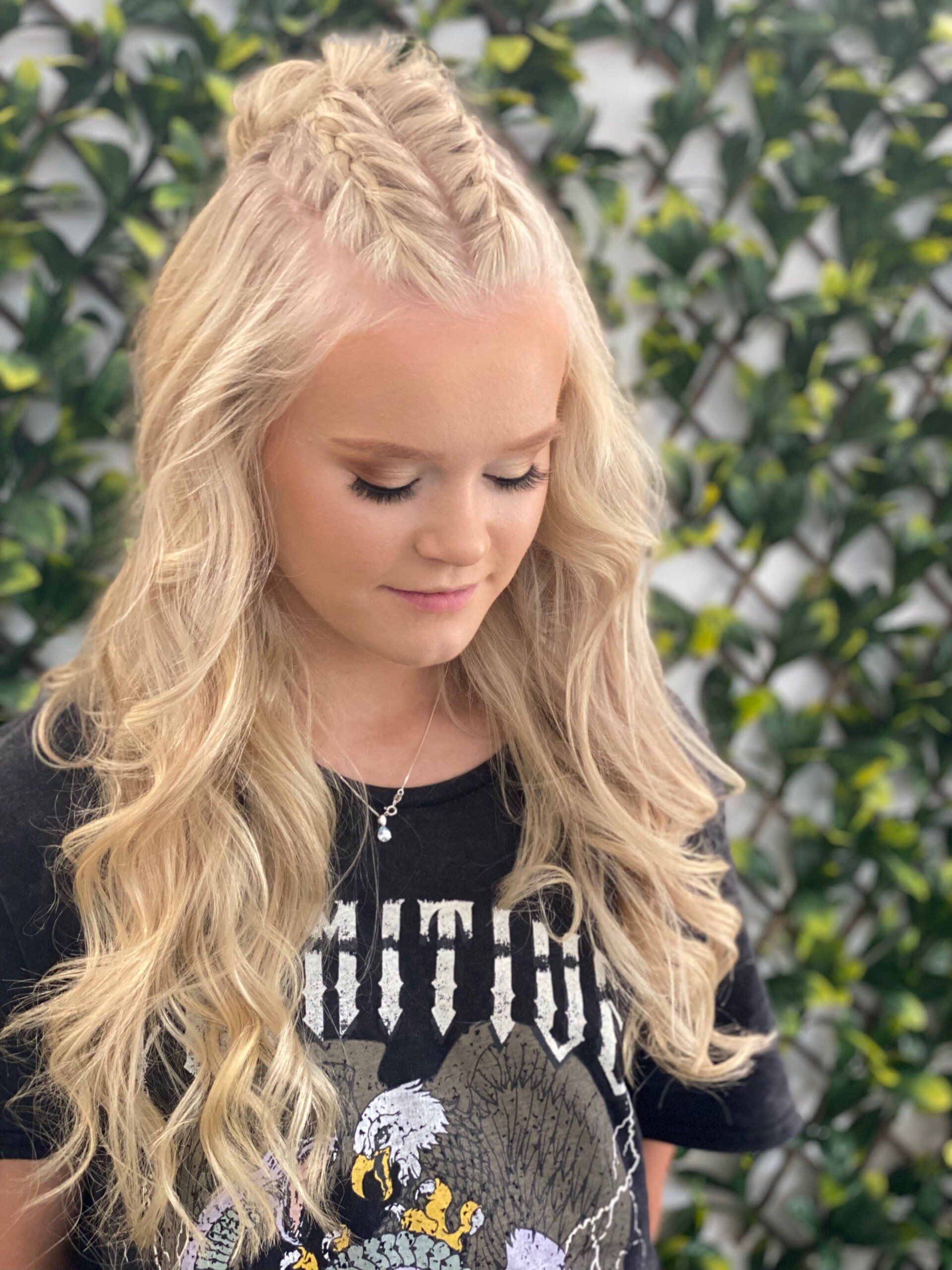 Blonde woman with half-up braided hairstyle, wearing a black t-shirt, outdoors with greenery background.