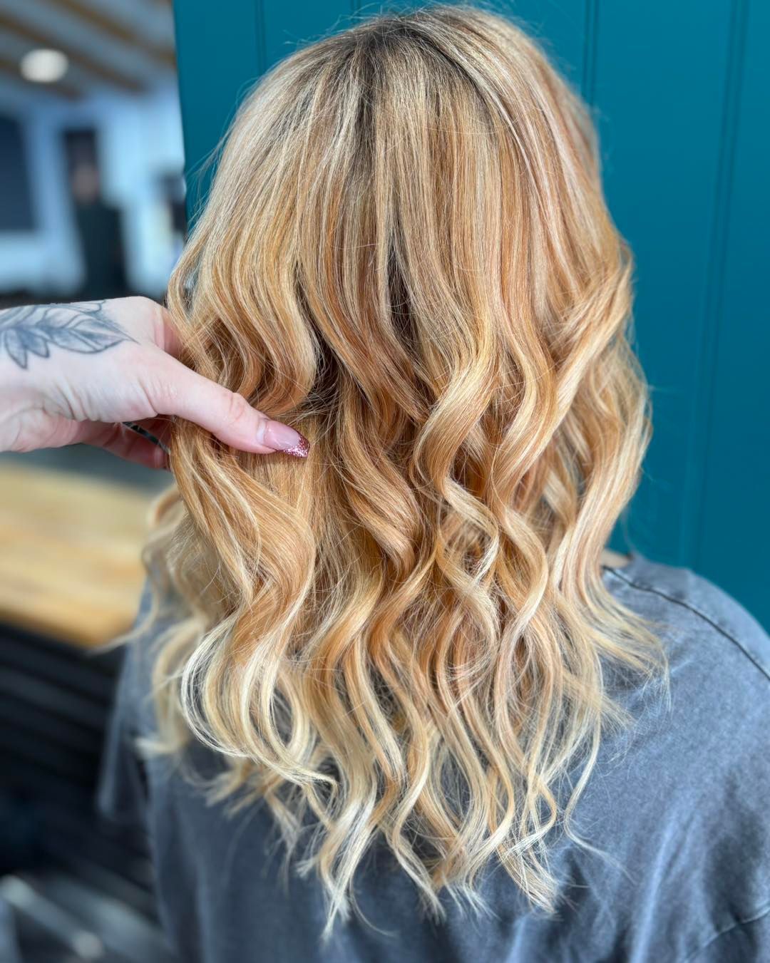Woman's wavy blonde hair with highlights, hand touching hair, teal wall background.