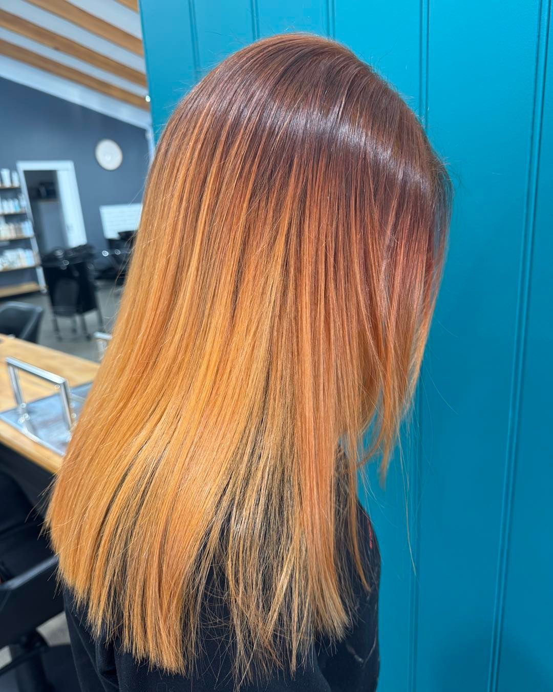 Woman with reddish-orange ombre hair, styled straight. Indoors, turquoise wall in background.