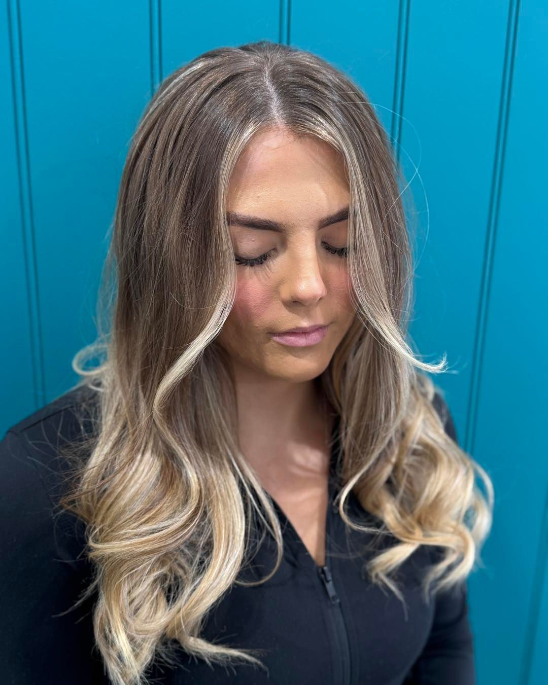 Woman with blonde highlights and loose waves, eyes closed, in front of a teal wall.