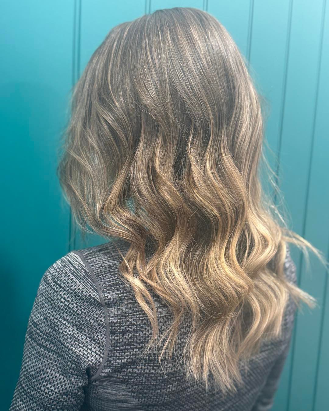 Woman with wavy, highlighted brown hair; standing in front of a teal wall.