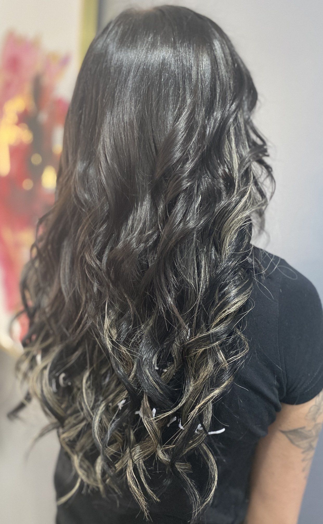 Woman with long, wavy, dark hair with blonde highlights, standing near a colorful wall.