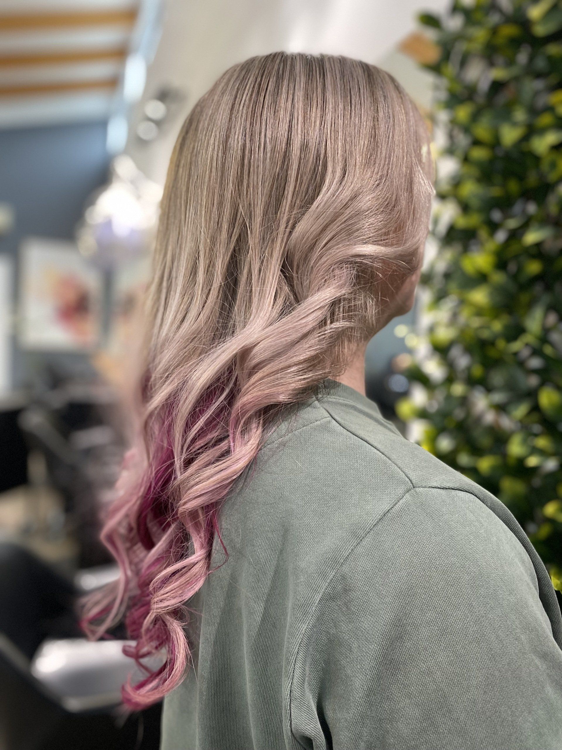 Woman with long, wavy, ombre hair, transitioning from light brown to pink. She wears a green sweater in a salon.