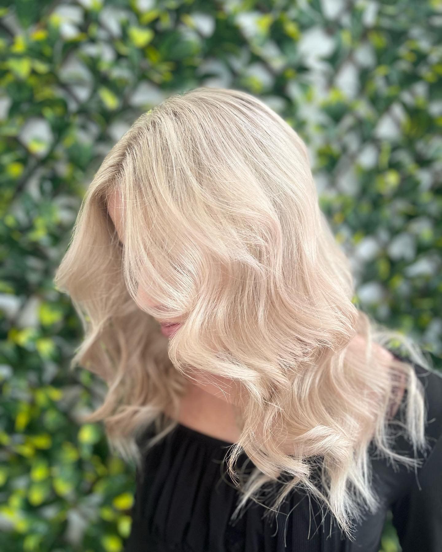 Woman with wavy, light blonde hair, against a green, leafy backdrop.
