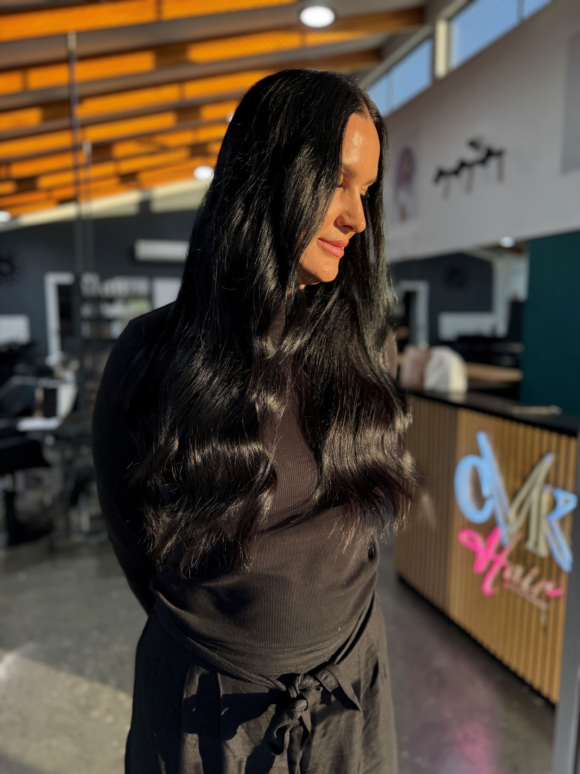 Person with long black hair in a black outfit standing indoors near a reception counter and warm-lit ceiling.