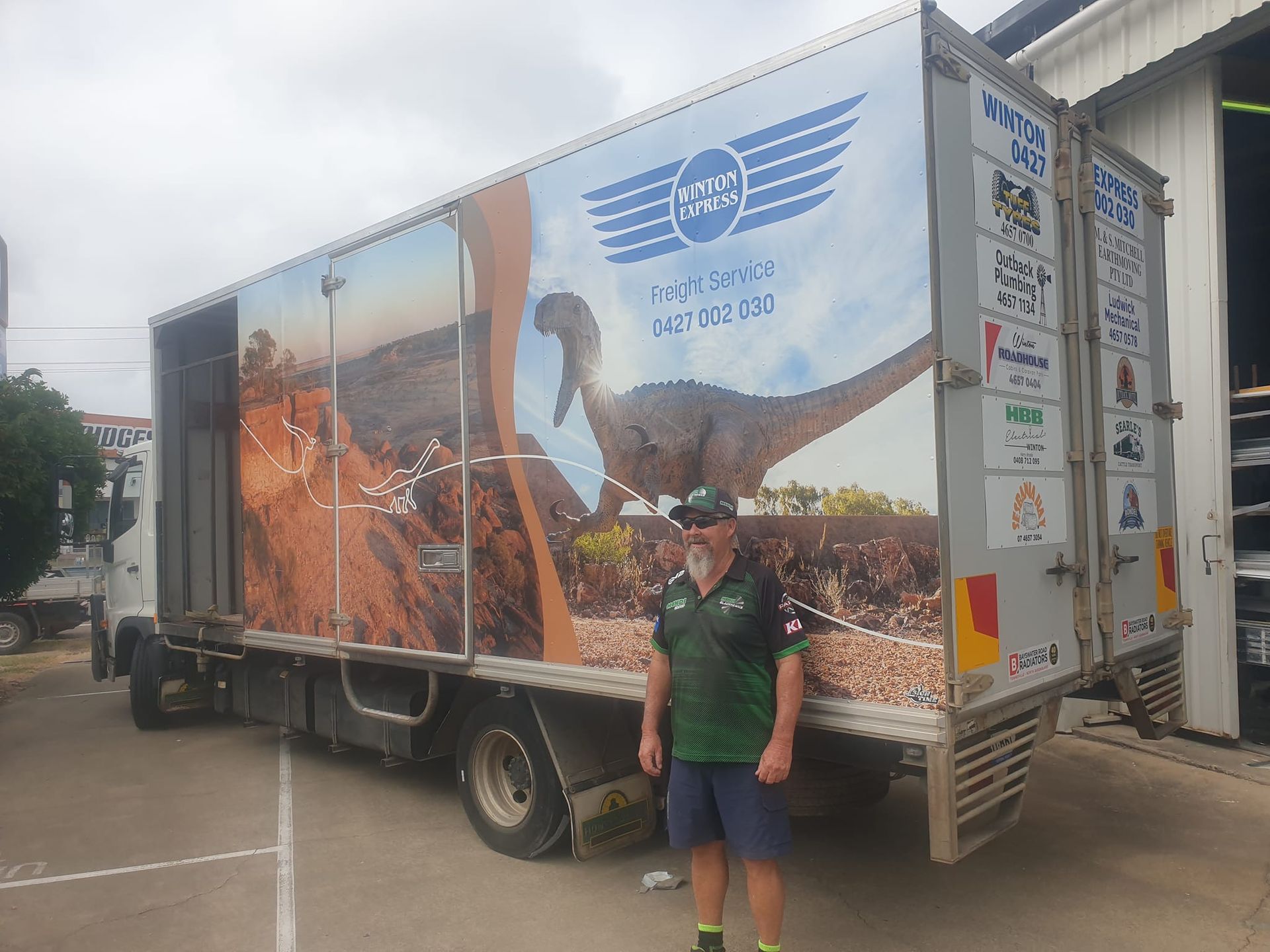 A man is standing in front of a large truck with a dinosaur on the side.