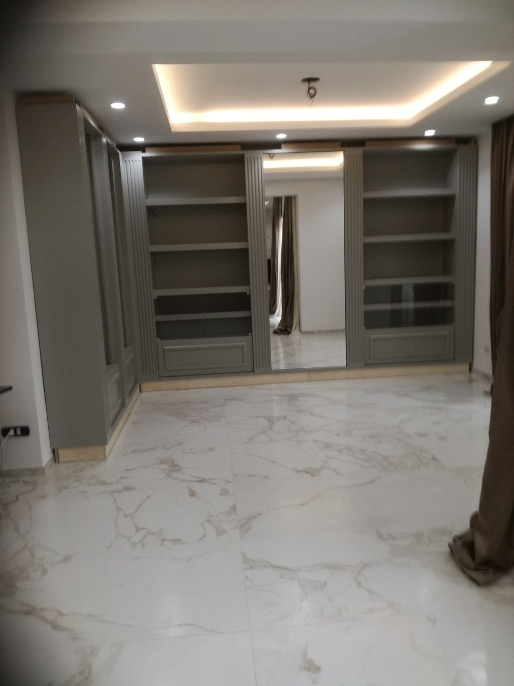 Cabina armadio moderna e vuota con scaffali a muro, specchio, illuminazione a incasso e pavimento effetto marmo.