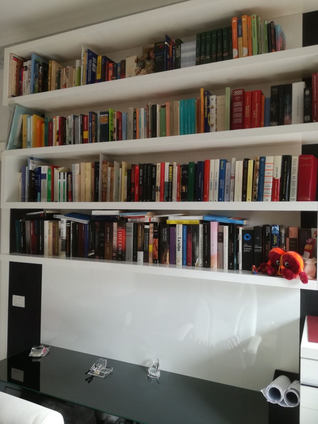 Scaffali pieni di libri sopra una scrivania nera, con un piccolo peluche e delle casse acustiche sulla destra.