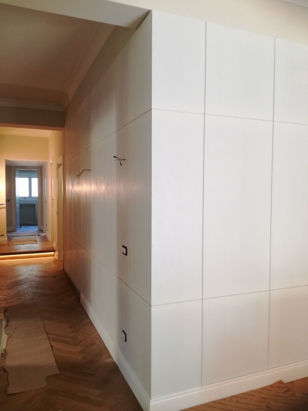 Stretto corridoio con armadi a muro bianchi e pavimento in legno in fase di ristrutturazione, che conduce a una luminosa porta d'ingresso.