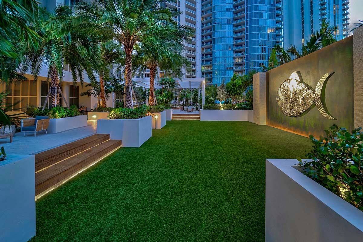 Modern apartment courtyard with green lawn, steps, palm trees, and a decorative wall at dusk