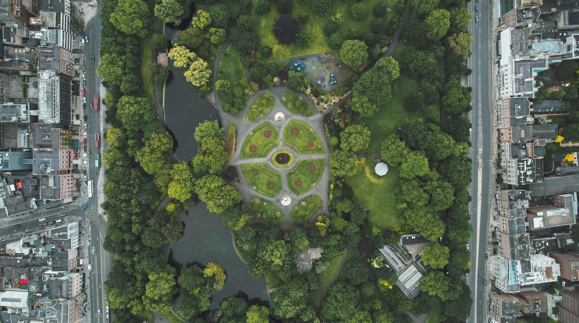 Aerial view of a park with trees, paths, and a pond, bordered by dense city buildings.