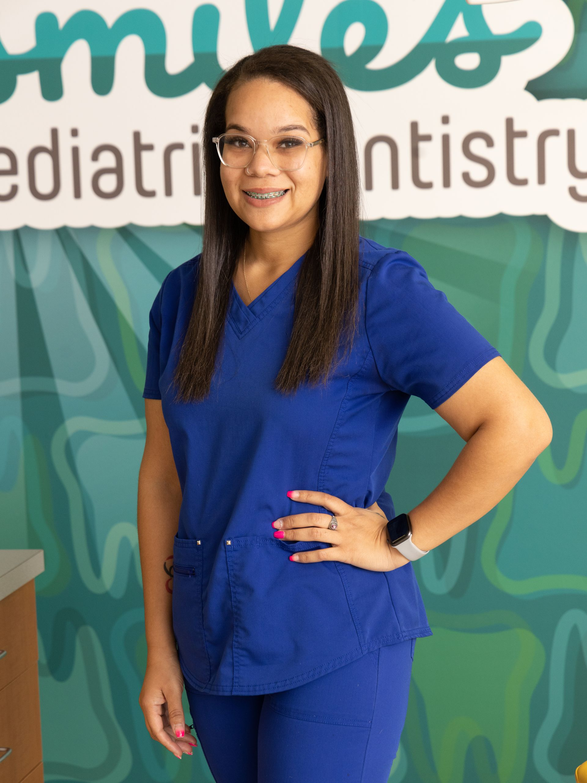 Woman in blue scrubs smiles at camera, standing in a pediatric dentistry office.
