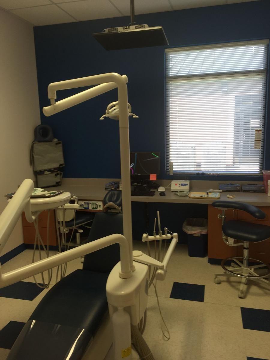 Dental examination room with blue chair, tools, overhead light, and a window with blinds.