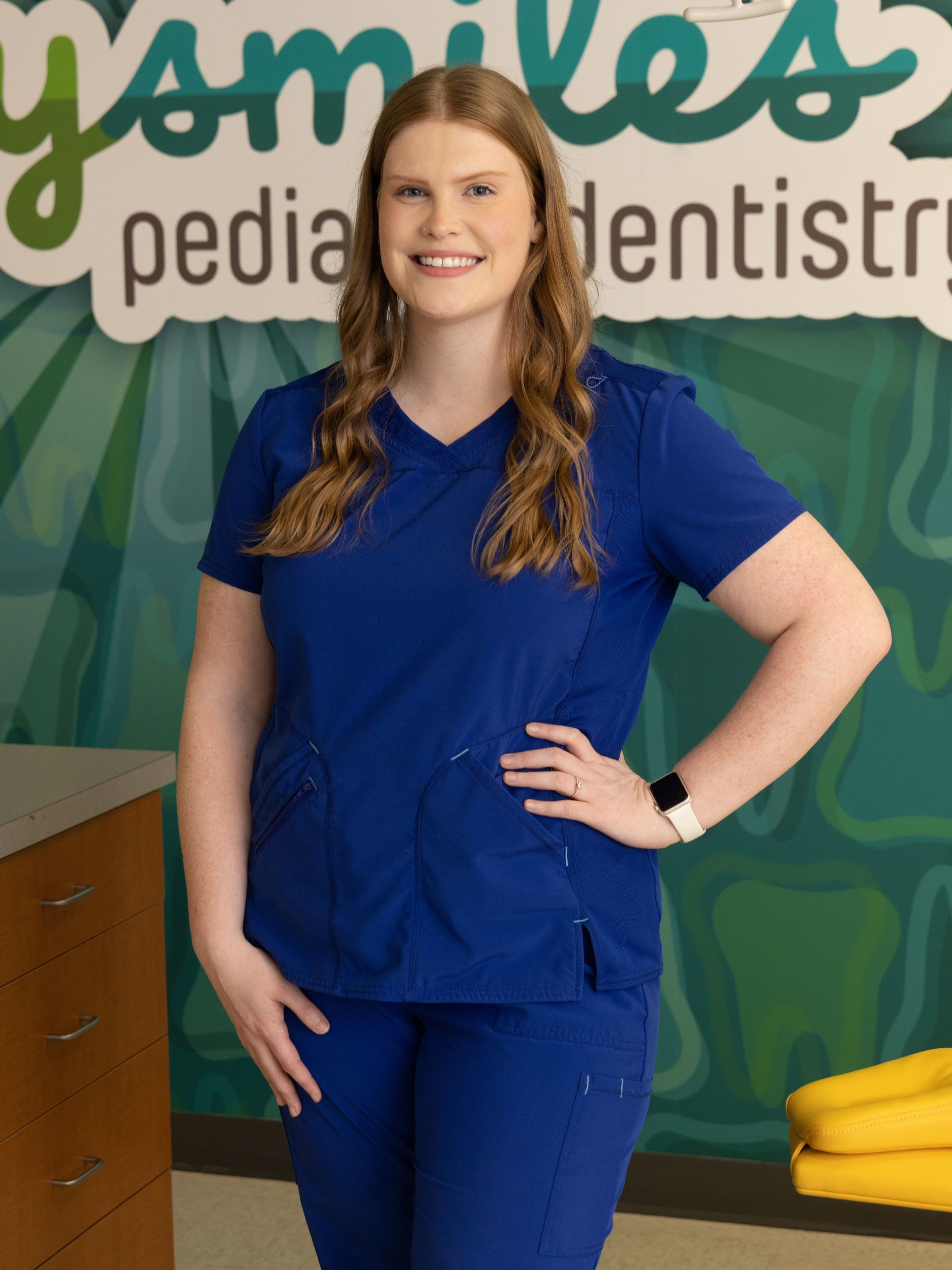 Woman in blue scrubs smiles in a pediatric dentistry office, hand on hip.