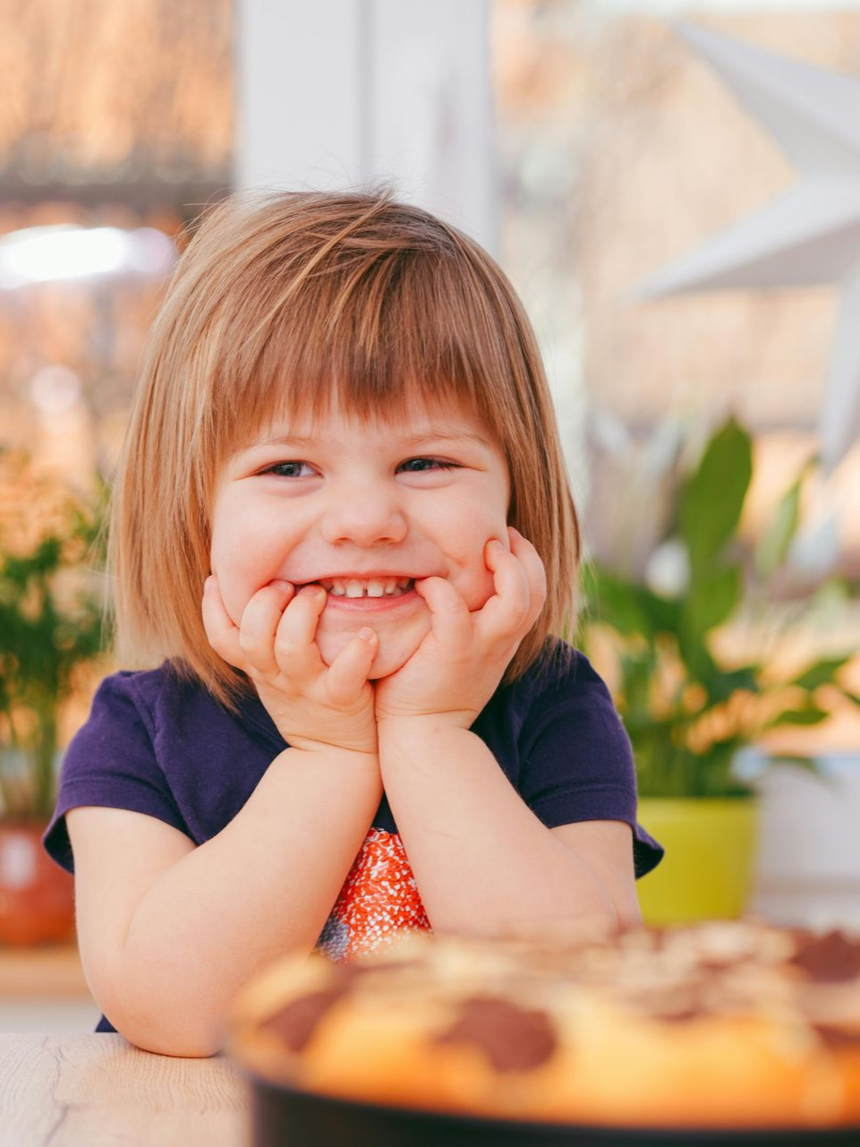 Young child smiling with hands under chin, looking at a cake.