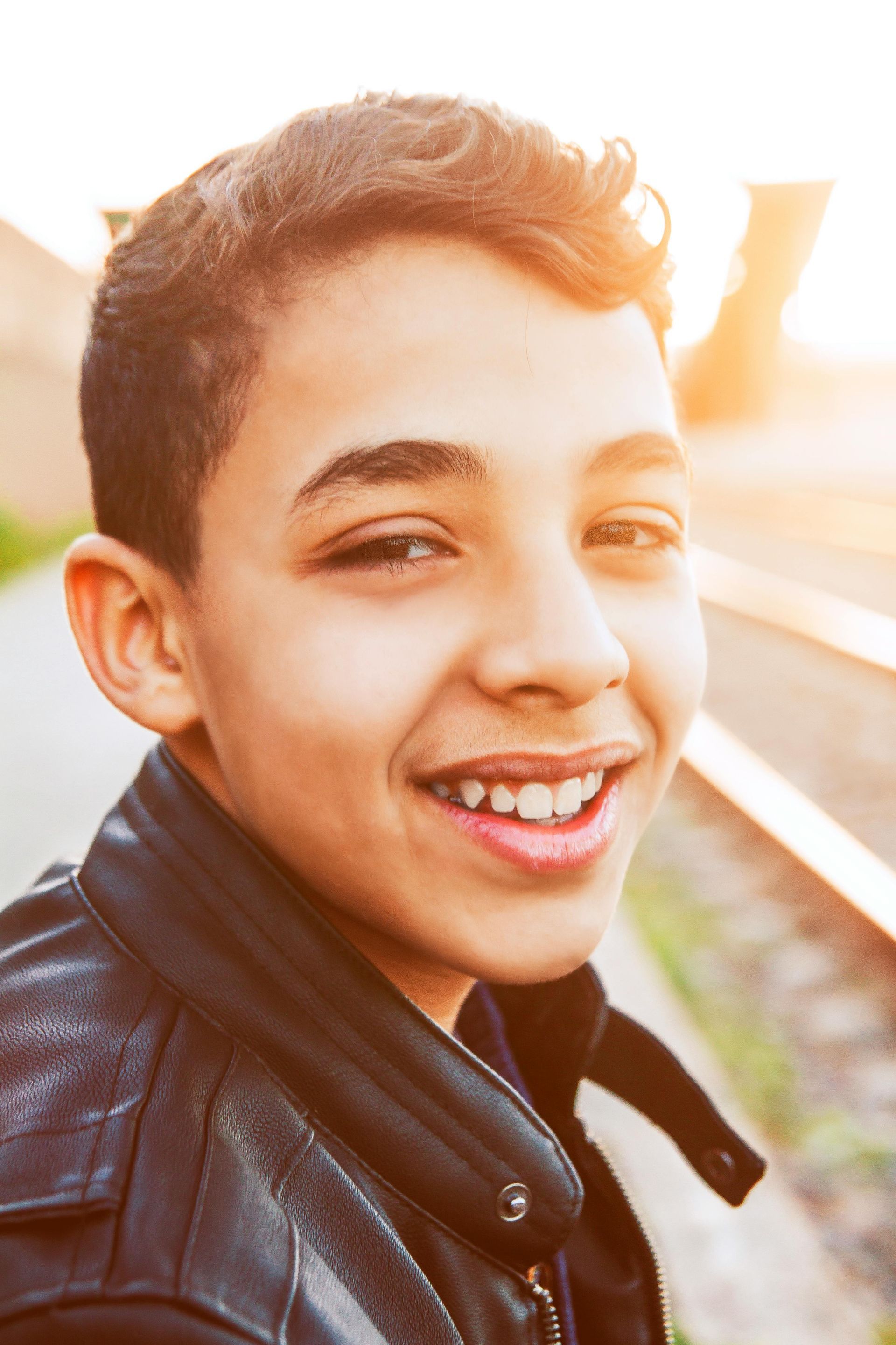 Boy smiling outdoors, wearing a black jacket, near train tracks at sunset.
