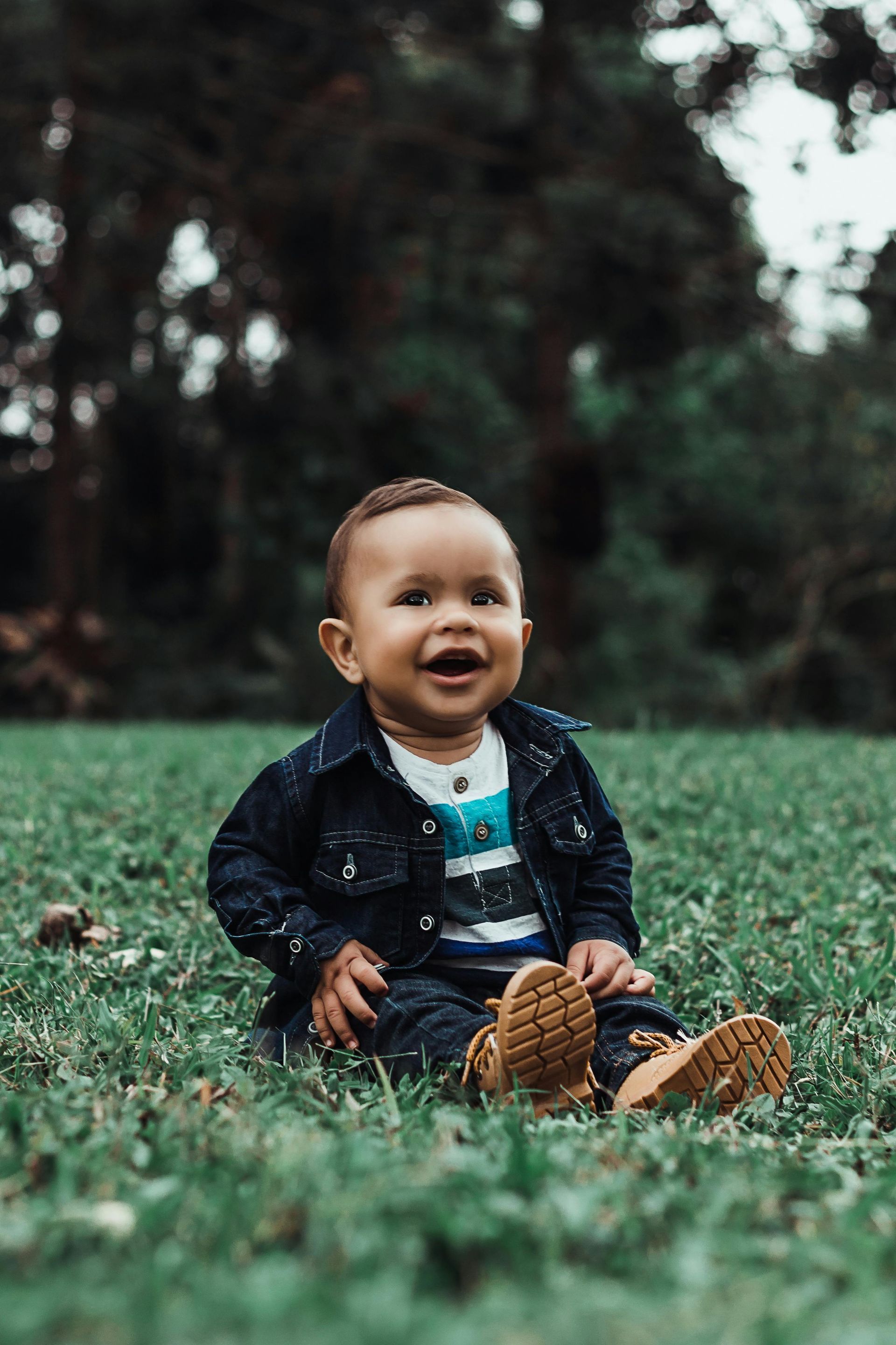 Smiling baby sits in grass wearing a denim jacket and boots, with a forest background.