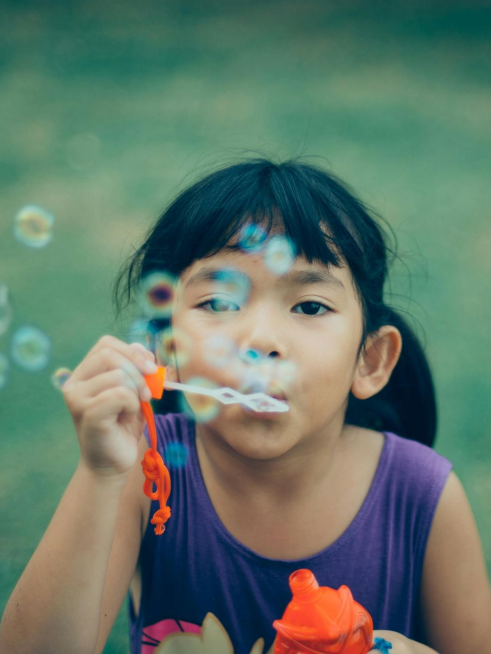 Girl blowing bubbles, wearing purple tank top, holding bubble wand, outdoors with blurred green background.