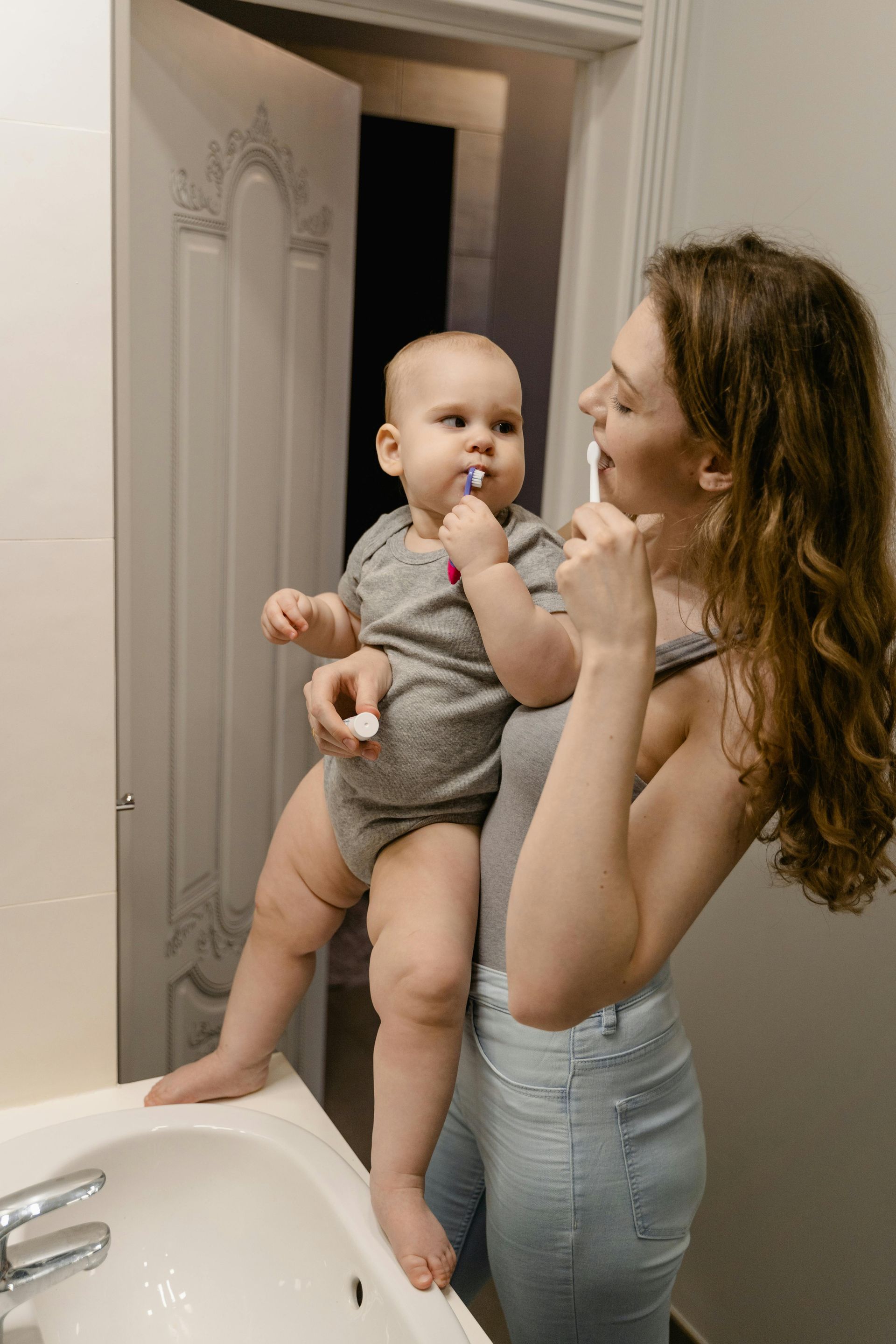 Woman brushing teeth with baby in a bathroom. Both hold toothbrushes.