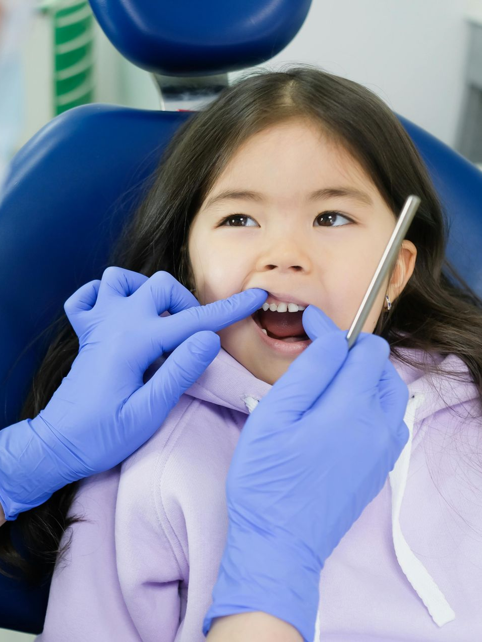 Girl in dental chair, mouth open, examined by gloved hands. Blue chair, dental tools.